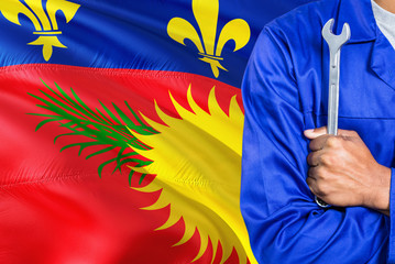 Mechanic in blue uniform is holding wrench against waving Guadeloupe flag background. Crossed arms technician.