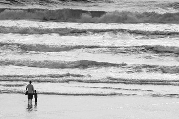 Fototapeta na wymiar Father and son holding hands on a pacific beach at sunset with waves in black and white.