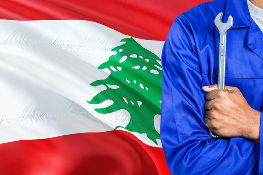 Lebanese Mechanic in blue uniform is holding wrench against waving Lebanon flag background. Crossed arms technician.