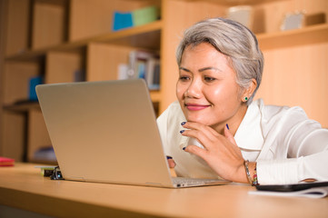 natural lifestyle office portrait of attractive and happy successful mature Asian woman working at laptop computer desk smiling confident in entrepreneur success