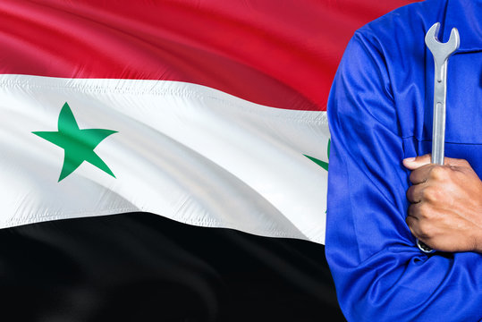 Syrian Mechanic in blue uniform is holding wrench against waving Syria flag background. Crossed arms technician.