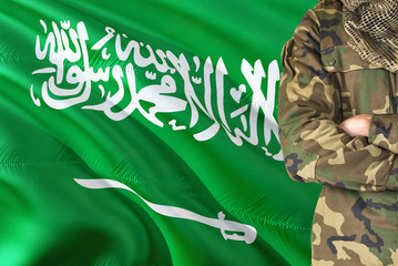 Fototapeta premium Crossed arms Saudi soldier with national waving flag on background - Saudi Arabia Military theme.