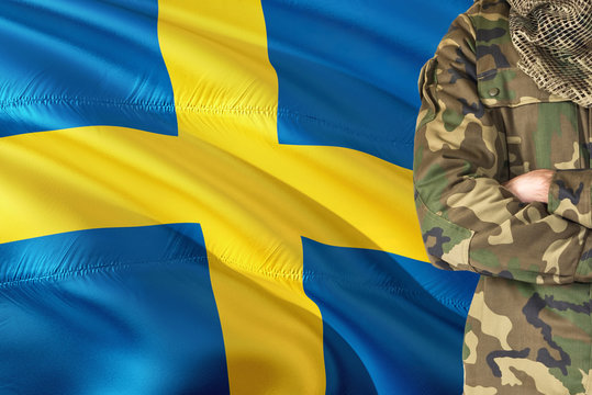 Crossed arms Swedish soldier with national waving flag on background - Sweden Military theme.