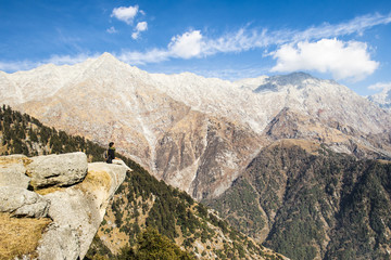 A tourist is sitting on a cliff enjoying the beautiful view of Dhauladhar Mountain ranges during a sunny day. Triund, Dharamsala, Himachal Pradesh, India.