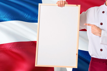 Dominican Chef holding blank whiteboard menu on Dominican Republic flag background. Cook wearing uniform pointing space for text.