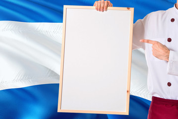Nicaraguan Chef holding blank whiteboard menu on Nicaragua flag background. Cook wearing uniform pointing space for text.