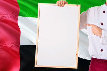 Chef holding blank whiteboard menu on United Arab Emirates flag background. Cook wearing uniform pointing space for text.