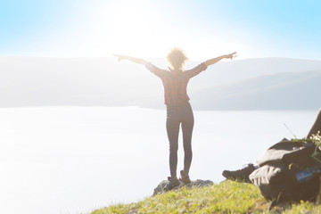 Young happy woman with backpack standing on a rock with raised hands and looking to a valley below