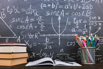 Books and stand for pens on a wooden table, against the background of a chalk board with formulas. Teacher's day concept and back to school.