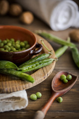 view of some raw fresh green peas in an earthenware plate and some pea pods on a brown rustic wooden table and linen