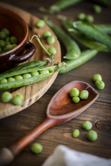 view of some raw fresh green peas in an earthenware plate and some pea pods on a brown rustic wooden table and linen