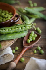 view of some raw fresh green peas in an earthenware plate and some pea pods on a brown rustic wooden table and linen