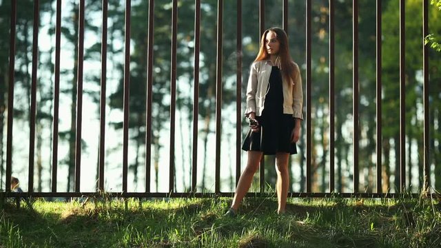 Vape teenager. Young cute girl in casual clothes smokes an electronic cigarette in front of a metal fence outdoors in the forest at sunset in summer. Bad habit. Stop vaping.