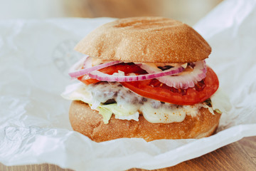 Delicious fresh homemade cheese burger with grilled meat, cheese, tomatoes, lettuce and onion rings in kraft paper. street food on wooden background