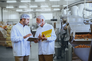 Two serious supervisors in sterile uniforms talking about quality of salty sticks. Older one...