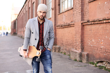 Handsome young man in grey coat and hat walking on the street, using longboard.