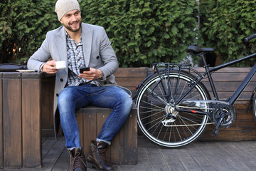 Obraz premium Handsome young man in grey coat and hat using smartphone, resting, and smiling while sitting near his bicycle outdoors.