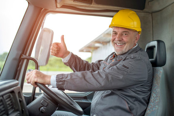 Smiling truck driver with hard hat inside the cabin showing thumbs up