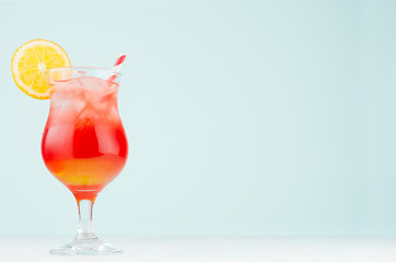 Bright layered red, yellow alcohol cocktail with oranges slice, straw, ice in elegant wineglass on soft light pastel blue background.