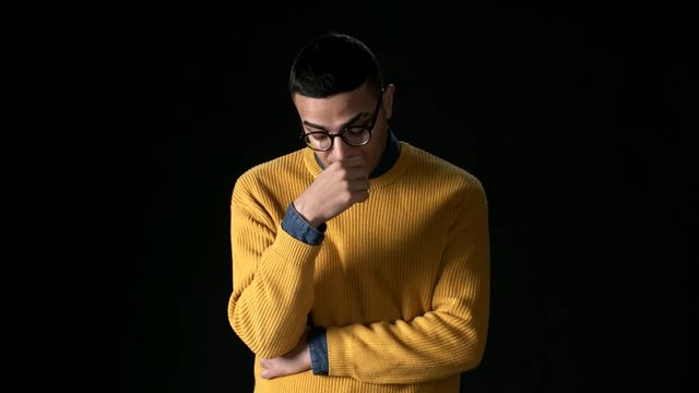 Medium shot of thoughtful young Arab man in spectacles and yellow sweater standing, touching his forehead with his hand then raising his head and looking at camera