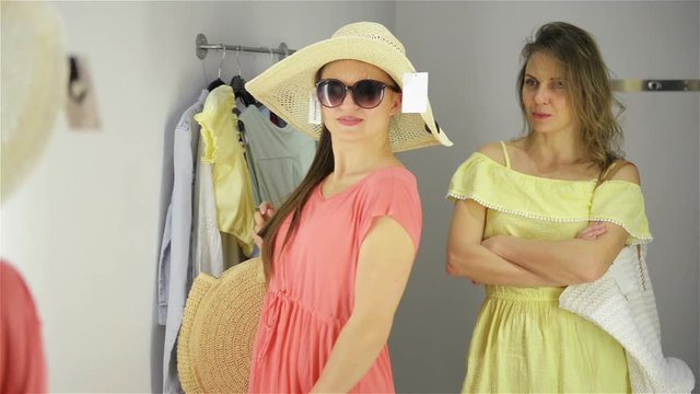 Two Women Shopping Trying A Funny Hat With Sunglasses. Beautiful Young Shoppers In Fitting Room.