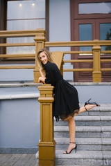 Young beautiful girl in a black dress is standing on the stairs, raised one leg and hand to the side. The concept of joy and carefree happiness of a young beautiful girl