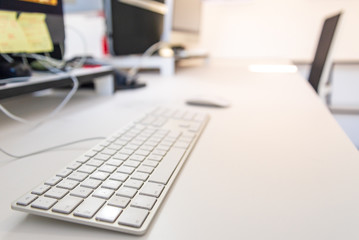 Closeup view of keyboard on white desktop, and blurry background of yellow notepads on desktop computer's monitors and office accessories with white interior atmosphere of real office working space.