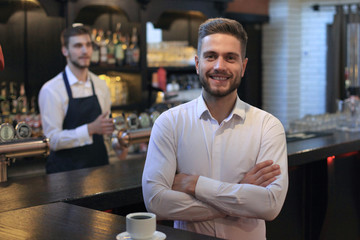 Successful small business owner standing with crossed arms with employee in background preparing...