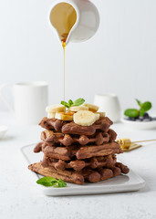 Chocolate banana waffles on white table, side view, vertical. Sweet brunch, maple syrup flow in milk jug, creamer.
