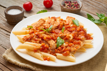 Penne pasta, chicken or turkey fillet, tomato sauce with basil leaves on old rustic wooden background.