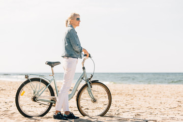 Fototapeta premium Pretty blonde girl in white pants and denim coat standing on the beach with bicycle. Atrractive woman relaxing near the sea after bike ride