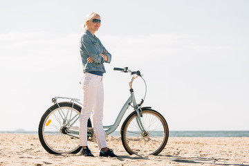 Fototapeta premium Pretty blonde girl in white pants and denim coat standing on the beach with bicycle. Atrractive woman relaxing near the sea after bike ride