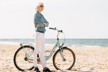 Fototapeta premium Pretty blonde girl in white pants and denim coat standing on the beach with bicycle. Atrractive woman relaxing near the sea after bike ride