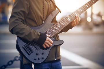 A man dressed in a dark jacket with striped laces plays a beautiful black five-string bass guitar on the street. - obrazy, fototapety, plakaty