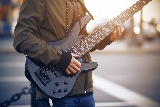 A man dressed in a dark jacket with striped laces plays a beautiful black five-string bass guitar on the street.
