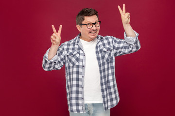 Naklejka premium Portrait of funny happy middle aged man in casual checkered shirt, eyeglasses standing with peace or victory sign and looking at camera with toothy smile. studio shot, isolated on dark red background.