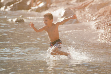 Fototapeta premium happy active little delightful boy in sport shorts running in sea water with splash on summer sunny beach
