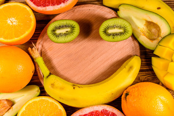 Still life with exotic fruits and cutting board. Bananas, mango, oranges, avocado, grapefruit and kiwi fruits on wooden table. Top view. Smiling face