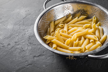 cooked Penne rigate pasta in stainless steel colander on Dark grey black slate background with copy...