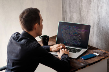 a male programmer in a black shirt is working at a laptop, close-up of the program code and developer's hands on a laptop. modern young programmer sitting at a wooden table at a computer