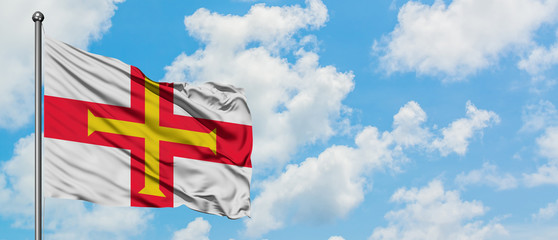 Guernsey flag waving in the wind against white cloudy blue sky. Diplomacy concept, international relations.