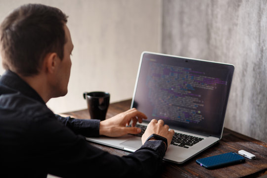 a male programmer in a black shirt is working at a laptop, close-up of the program code and developer's hands on a laptop. modern young programmer sitting at a wooden table at a computer