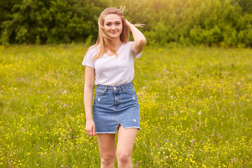 Naklejka na ściany i meble Outdoor shot of pretty blond woman wearing white casual t shirt and denim skirt, posing in meadow, touching her fair hair, looking at camera with pleasant facial exprassion. Happyness concept.