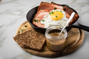 Gebräunter Bayrischer Fleischkäse mit gebratenen Spiegelei in Gusseisenpfanne mit süßen Senf und Brot auf Holzbrett