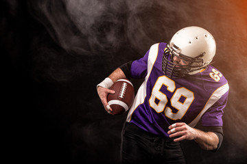 American football player holding ball in his hands in smoke. Black background, copy space. The concept of American football, motivation, copy space