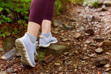 Outdoor shot of slender female legs in maroon leggings and grey sneakers go along forest trail. Walks in open fresh air, has jogging in mountainous terrain, having rest in green forest