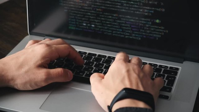 a male programmer in a black shirt is working at a laptop, close-up of the program code and developer's hands on a laptop. modern young programmer sitting at a wooden table at a computer