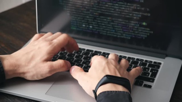 a male programmer in a black shirt is working at a laptop, close-up of the program code and developer's hands on a laptop. modern young programmer sitting at a wooden table at a computer