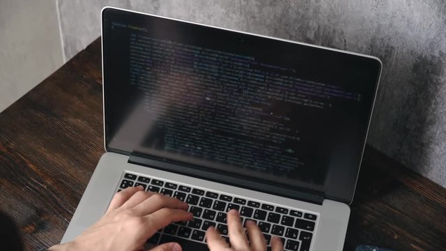 a male programmer in a black shirt is working at a laptop, close-up of the program code and developer's hands on a laptop. modern young programmer sitting at a wooden table at a computer