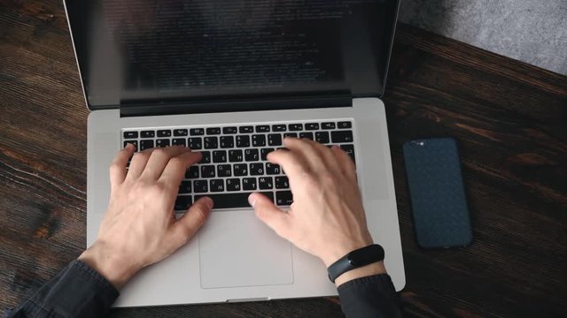 a male programmer in a black shirt is working at a laptop, close-up of the program code and developer's hands on a laptop. modern young programmer sitting at a wooden table at a computer